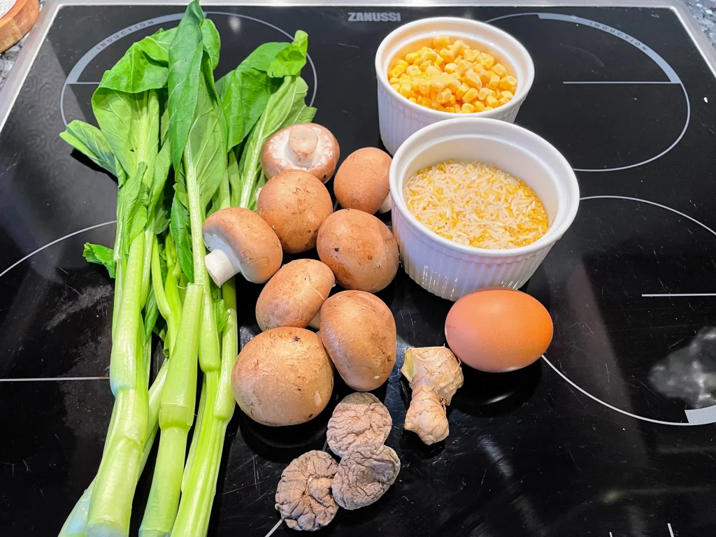 Ingredients for Chinese veggie congee laid out on a countertop, including rice, sweet corn, eggs, dried shiitake mushrooms, fresh chestnut mushrooms, ginger, leafy greens.